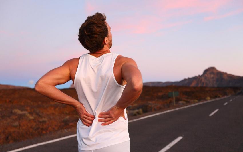 Back pain. Athletic running man with injury in sportswear rubbing touching lower back muscles standing on road outside at night.