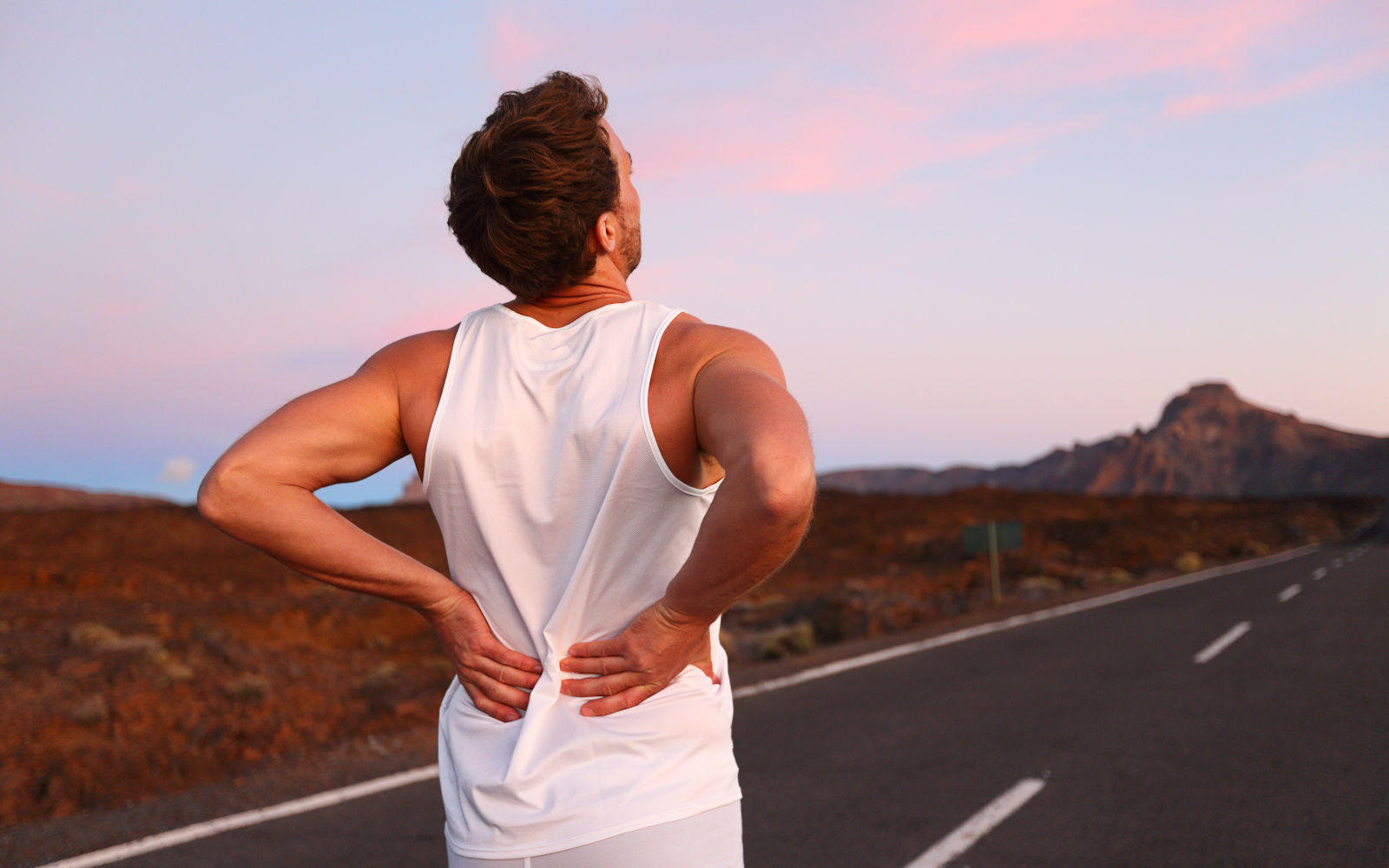 Back pain. Athletic running man with injury in sportswear rubbing touching lower back muscles standing on road outside at night.